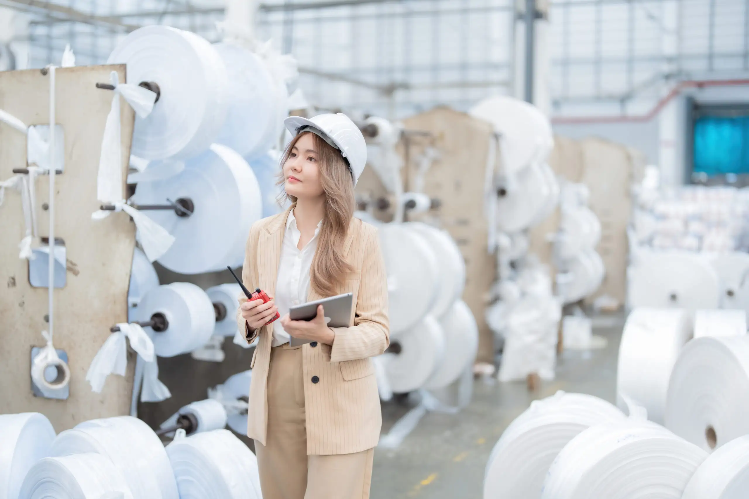 An Asian female engineer inspects a paper roll production machine in an industrial factory in preparation for export to a global customer. Wears a suit, hard hat, and holds a tablet and radio.